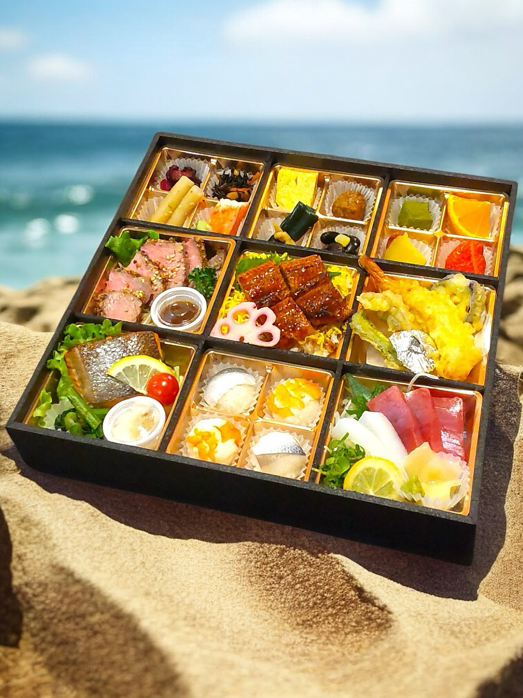 松山市の仕出し懐石弁当「花響」を、青空と海を背景に砂浜の上で撮影。色鮮やかな和食料理が並ぶ豪華な一折。
