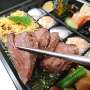 箸で持ち上げた、冷めてもやわらかい牛リブステーキ。彩り豊かな仕出し弁当の一品。