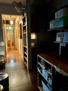 雨の朝、静かな仕込み場の通路。やわらかな灯りと木の香りが満ちる。